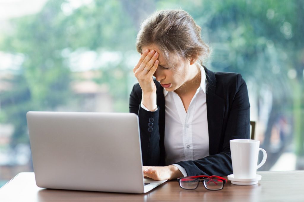 Young businesswoman sitting at table in front of laptop while closing eyes tight and holding hand on forehead. Front view with big window and blurry green view outside in background.