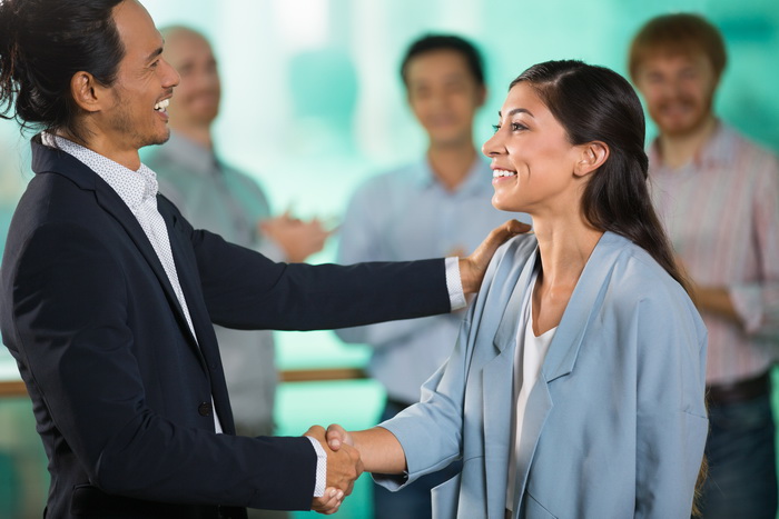 Smiling middle-aged Asian business man and Indian female colleague shaking hands and celebrating promotion with three blurred applauding people in background. Side view.