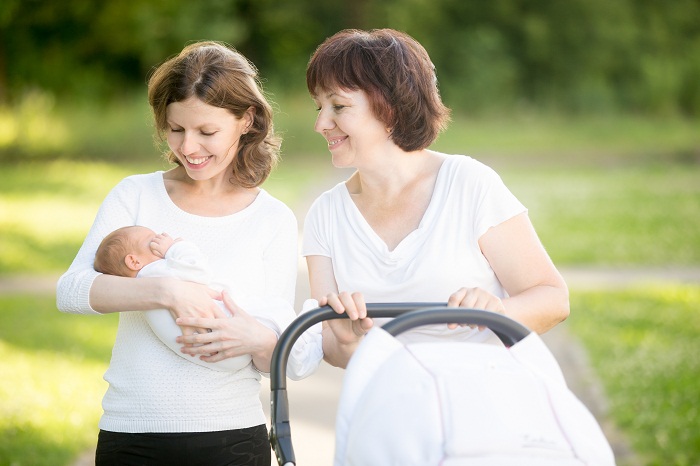 Portrait of three generations walking in park. Young pretty happy mother holding her new-born baby tightly on arms looking at it with love smiling. Grandma standing with pram looking at baby smiling